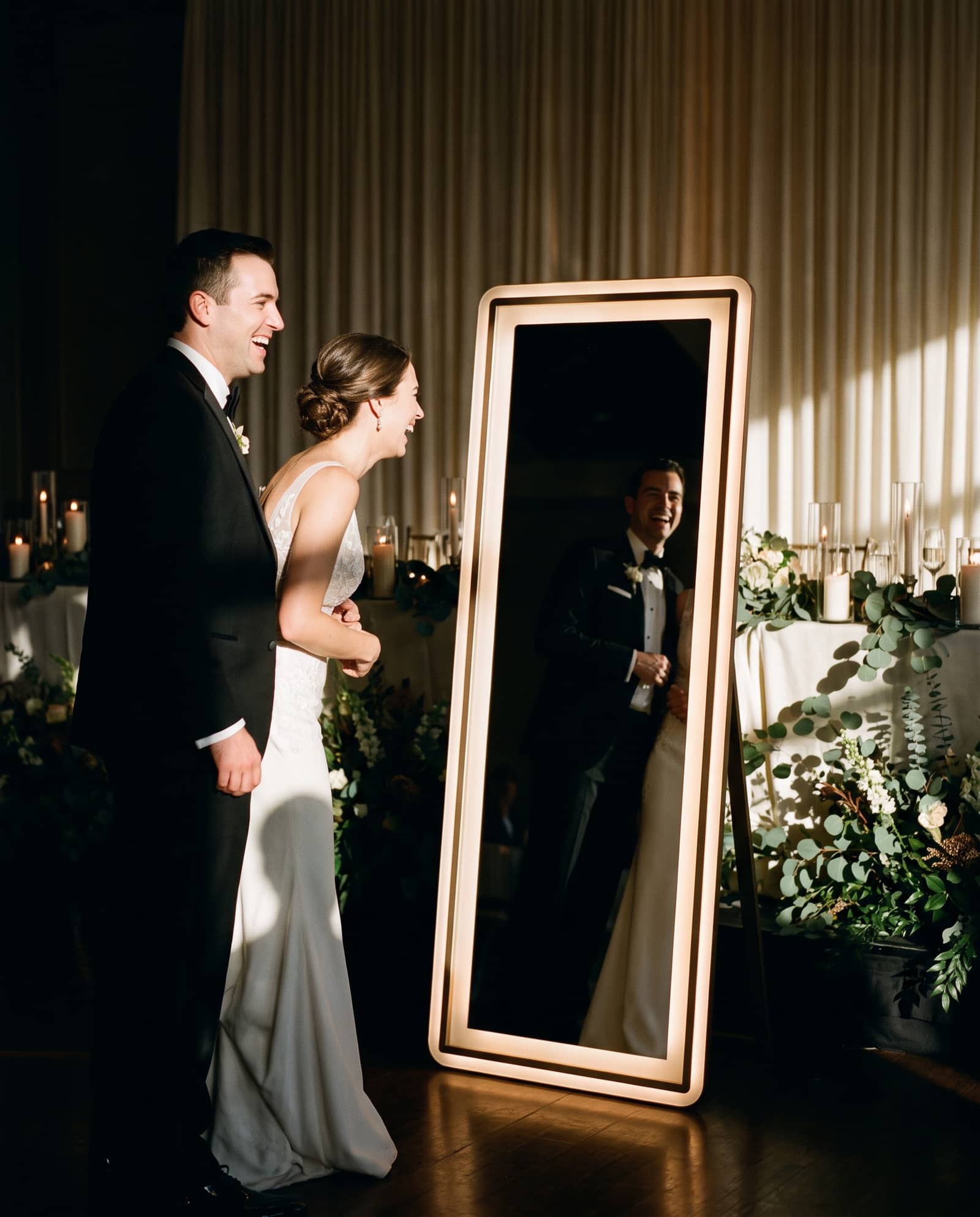 A bride and groom laughing in front of a full length magic mirror photo booth with a warm illuminated border at a candlelit reception