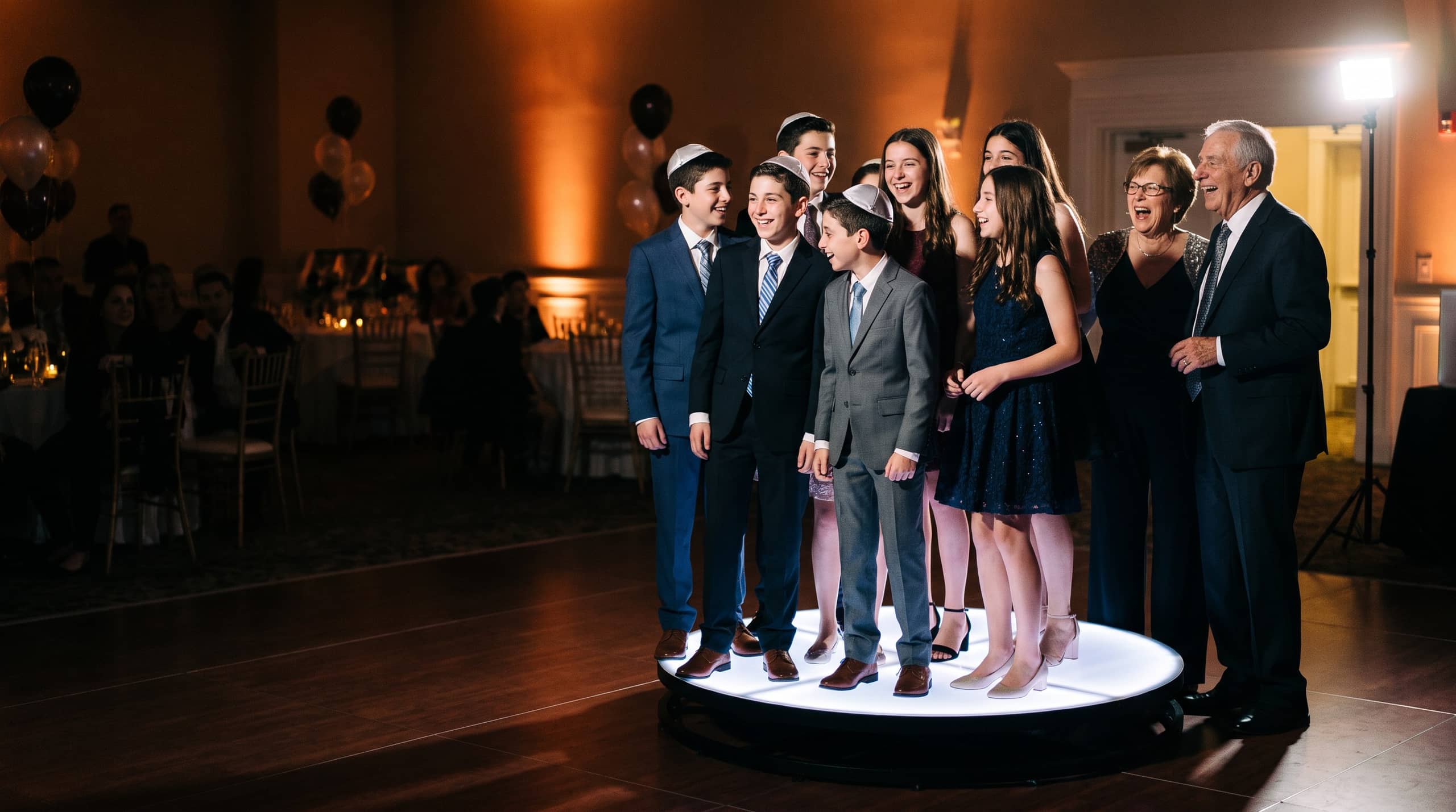 Teenagers in suits and party dresses laughing together on a lit 360 photo booth platform at a bar mitzvah reception, several of the boys wearing kippot, with grandparents laughing beside them